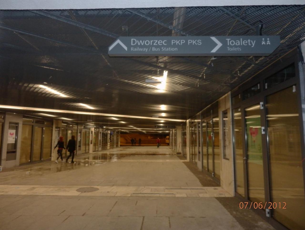 Podwieszany sufit z metalowej siatki w przejściu podziemnym z oświetleniem liniowym, widoczne znaki informacyjne: Dworzec PKP PKS oraz Toalety.