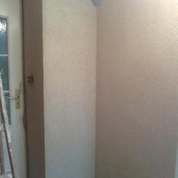 NAD-TEK usługi remontowo- budowlane - Ściana pokryta tynkiem strukturalnym typu 'Marmolit' w rogu pomieszczenia, widoczna drabina oparta o białe drzwi z szybkami.