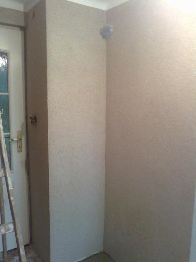 Ściana pokryta tynkiem strukturalnym typu 'Marmolit' w rogu pomieszczenia, widoczna drabina oparta o białe drzwi z szybkami.