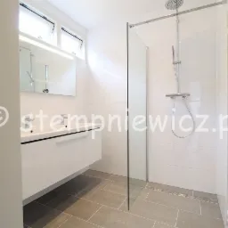 Nowoczesna łazienka z kabiną prysznicową typu walk-in, białą armaturą i szarą podłogą z płytek ceramicznych, widok na umywalkę z szafką i grzejnik drabinkowy.