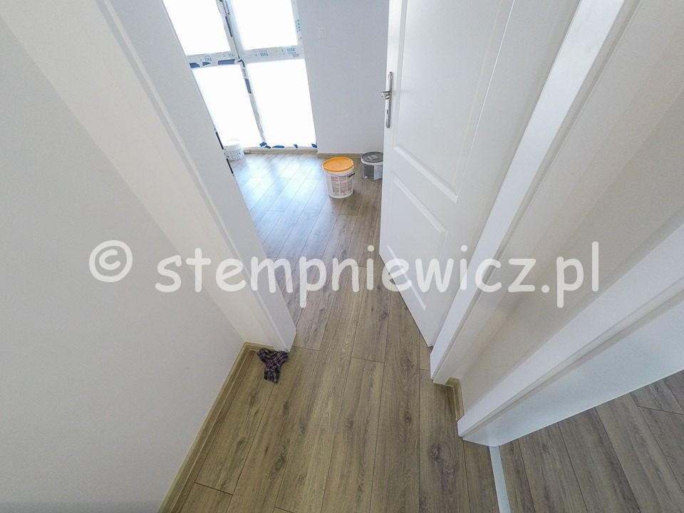 Wnętrze po remoncie: świeżo pomalowane białe ściany, jasna drewnopodobna podłoga, otwarte białe drzwi, widoczne pojemniki z farbą w tle przy oknie.