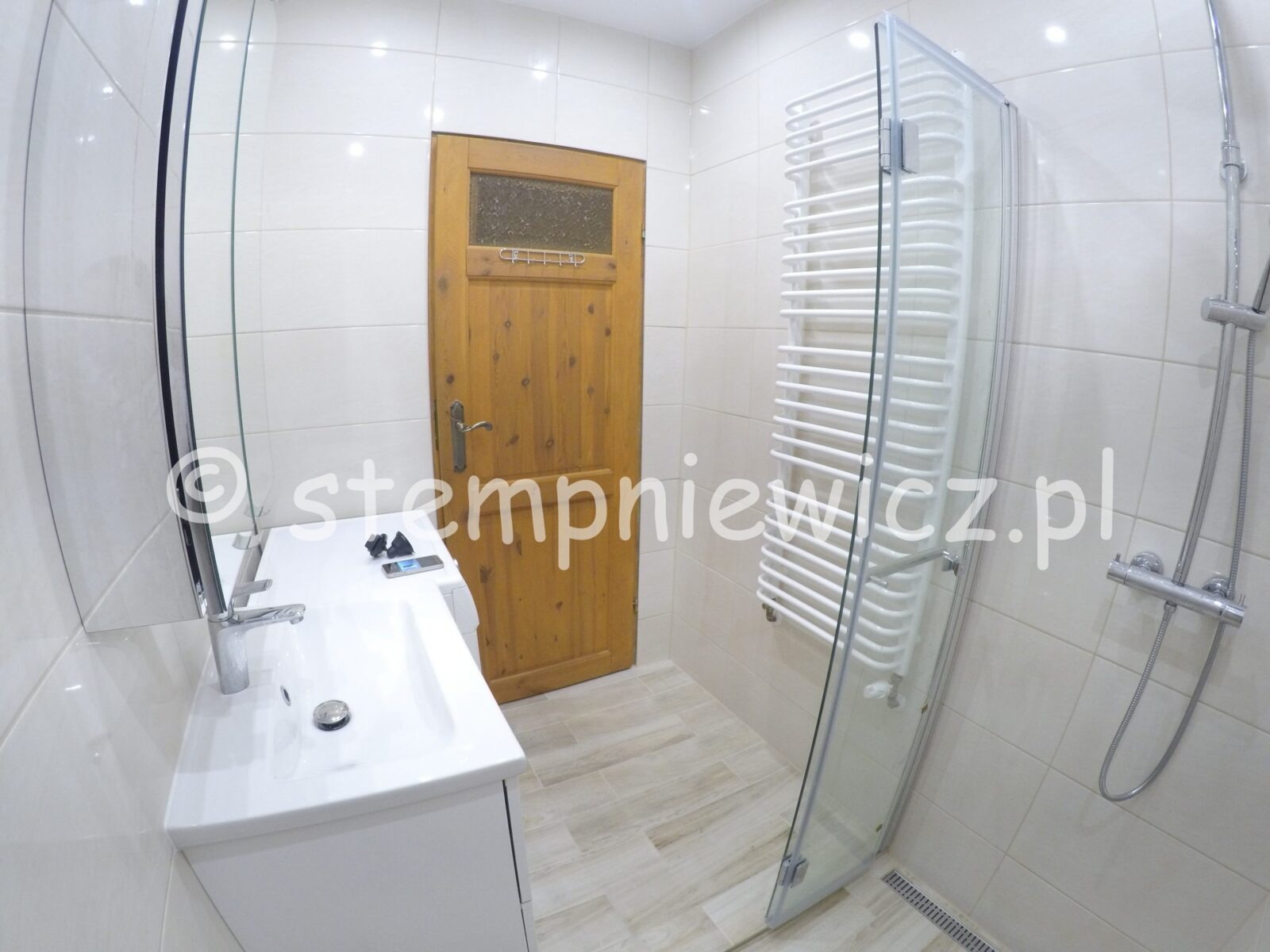 Jasna łazienka po remoncie z drewnianymi drzwiami, białą umywalką z szafką, grzejnikiem drabinkowym za szklaną ścianką prysznicową i chromowaną baterią prysznicową.