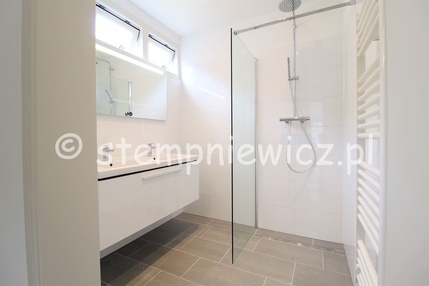 Nowoczesna łazienka z kabiną prysznicową typu walk-in, białą armaturą i szarą podłogą z płytek ceramicznych, widok na umywalkę z szafką i grzejnik drabinkowy.