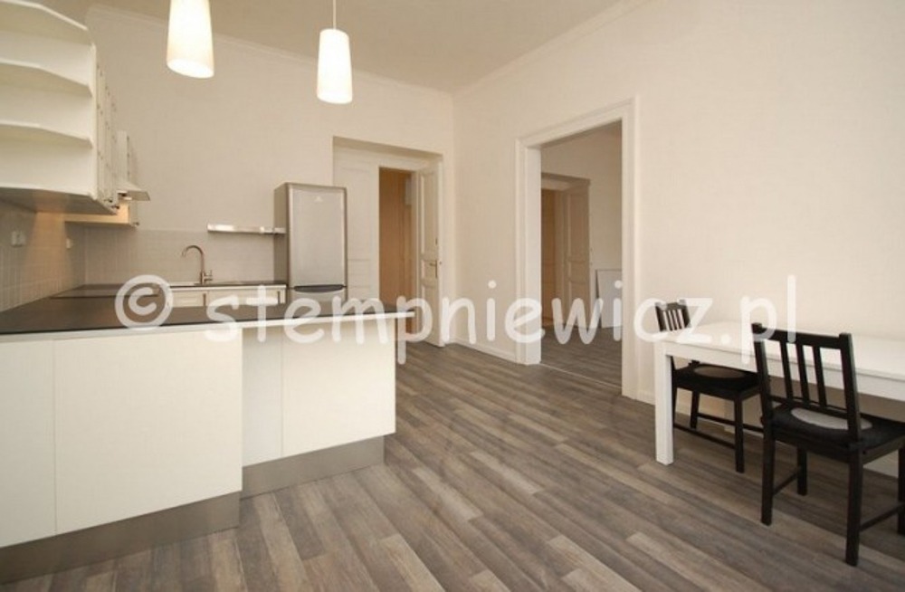 Jasne, przestronne wnętrze mieszkania z widoczną kuchnią z białymi szafkami i czarnym blatem, drewnopodobną podłogą oraz częścią jadalną z białym stołem i czarnymi krzesłami. W tle widoczne otwarte...