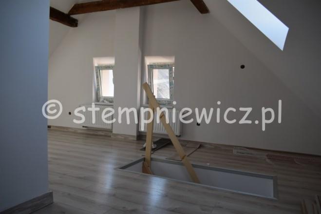 Poddasze z drewnianymi belkami, oknami połaciowymi i lukarnami. Widoczny otwór w podłodze z drewnianą konstrukcją wspornikową oraz fragment ułożonej podłogi z paneli. Na ścianie widoczny znak wodny.