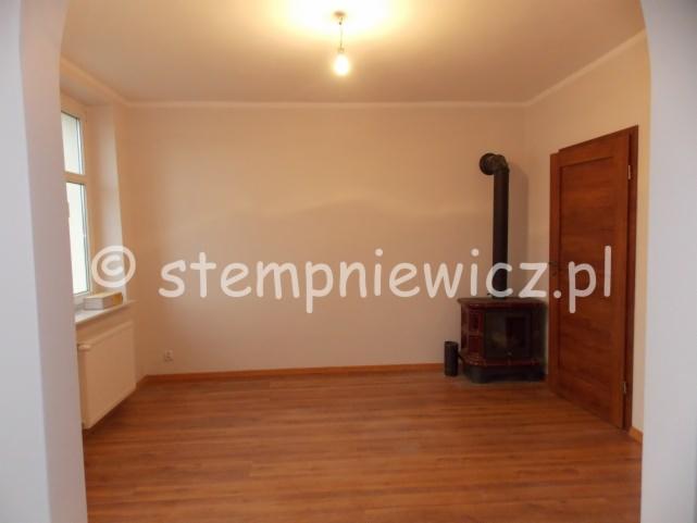 Pusty pokój z drewnianą podłogą, białymi ścianami i piecem wolnostojącym z czarną rurą odprowadzającą dym, widok na okno z grzejnikiem pod parapetem,  w prawym rogu drewniane drzwi.