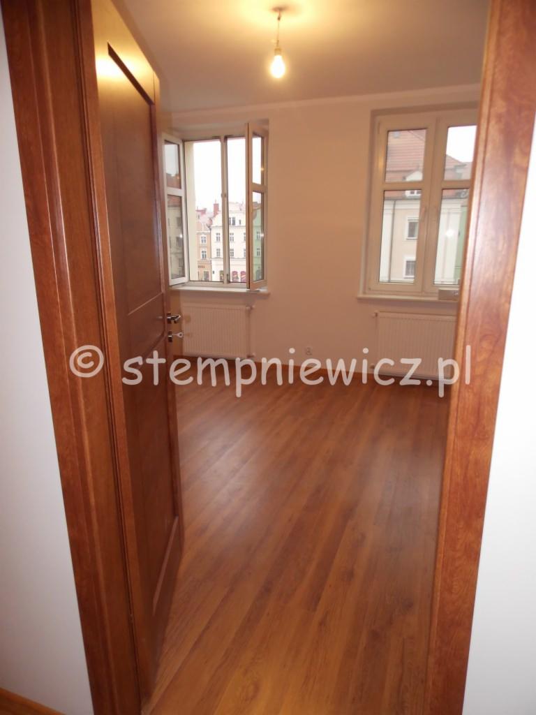 Widok na pokój z drewnianą podłogą ułożoną w deski, przez otwarte drzwi z drewnianą ościeżnicą, z oknami wychodzącymi na budynki za oknem.