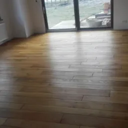 Ułożony parkiet z jasnego drewna w nowym, jasnym pokoju z dużym oknem balkonowym, widok na zewnątrz na plac budowy.