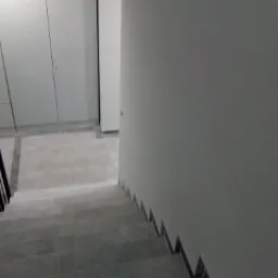 Schody wykończone szarymi płytkami prowadzące w dół, z widoczną białą ścianą i fragmentem białej szafy w tle.