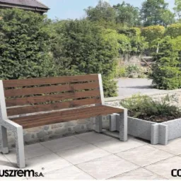 Drewniana ławka z betonowymi nogami i donice z betonu architektonicznego na tarasie z jasnych płyt, w tle zielony ogród.