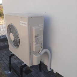 P.H.WASSER MANN II Alina Czapracka-Bodus - Zewnętrzna jednostka pompy ciepła marki De Dietrich, zamontowana na czarnej, metalowej konstrukcji wsporczej na kostce brukowej, z widocznym białym przewodem przyłączeniowym do budynku o białej...