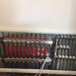 Rozdzielacz ogrzewania podłogowego z rurami w kolorach czerwonym i niebieskim, oznaczony etykietami dla poszczególnych pomieszczeń: łazienka mała, kuchnia, wiatrołap, salon, pokój gościnny...