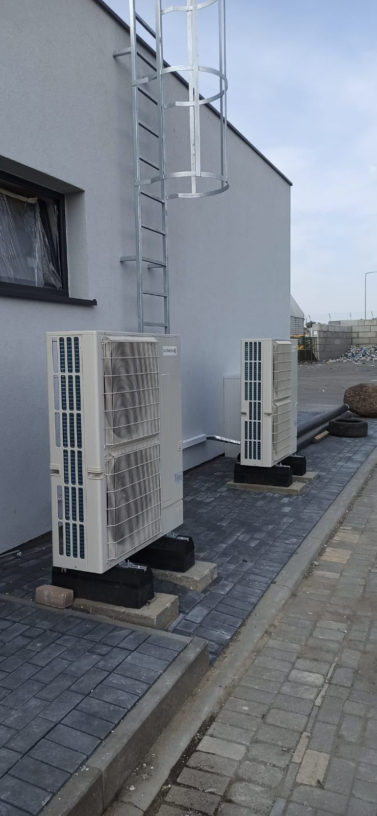 Dwa zewnętrzne moduły klimatyzacji De Dietrich ustawione na betonowych podstawach przy ścianie budynku z drabinką ewakuacyjną, widoczne przewody i kostka brukowa.