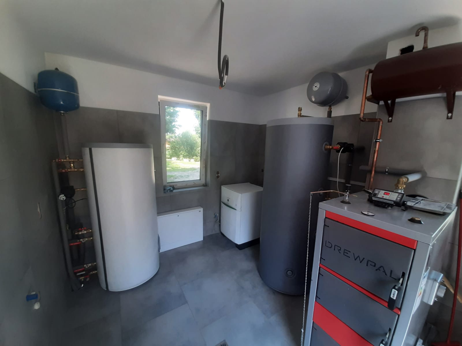 Kotłownia z zainstalowanym kotłem Drewpal, dwoma zbiornikami akumulacyjnymi ciepła, naczyniem wzbiorczym, grzejnikiem oraz oknem z widokiem na zieleń.