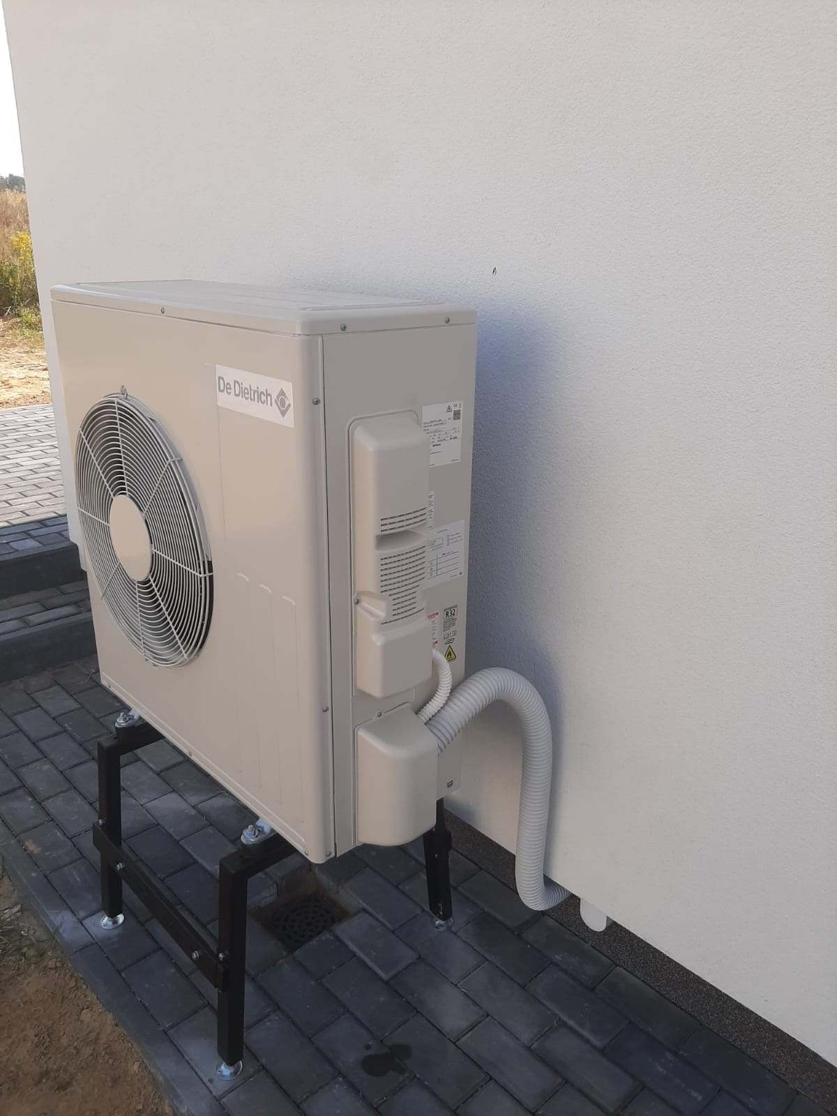 Zewnętrzna jednostka pompy ciepła marki De Dietrich, zamontowana na czarnej, metalowej konstrukcji wsporczej na kostce brukowej, z widocznym białym przewodem przyłączeniowym do budynku o białej...