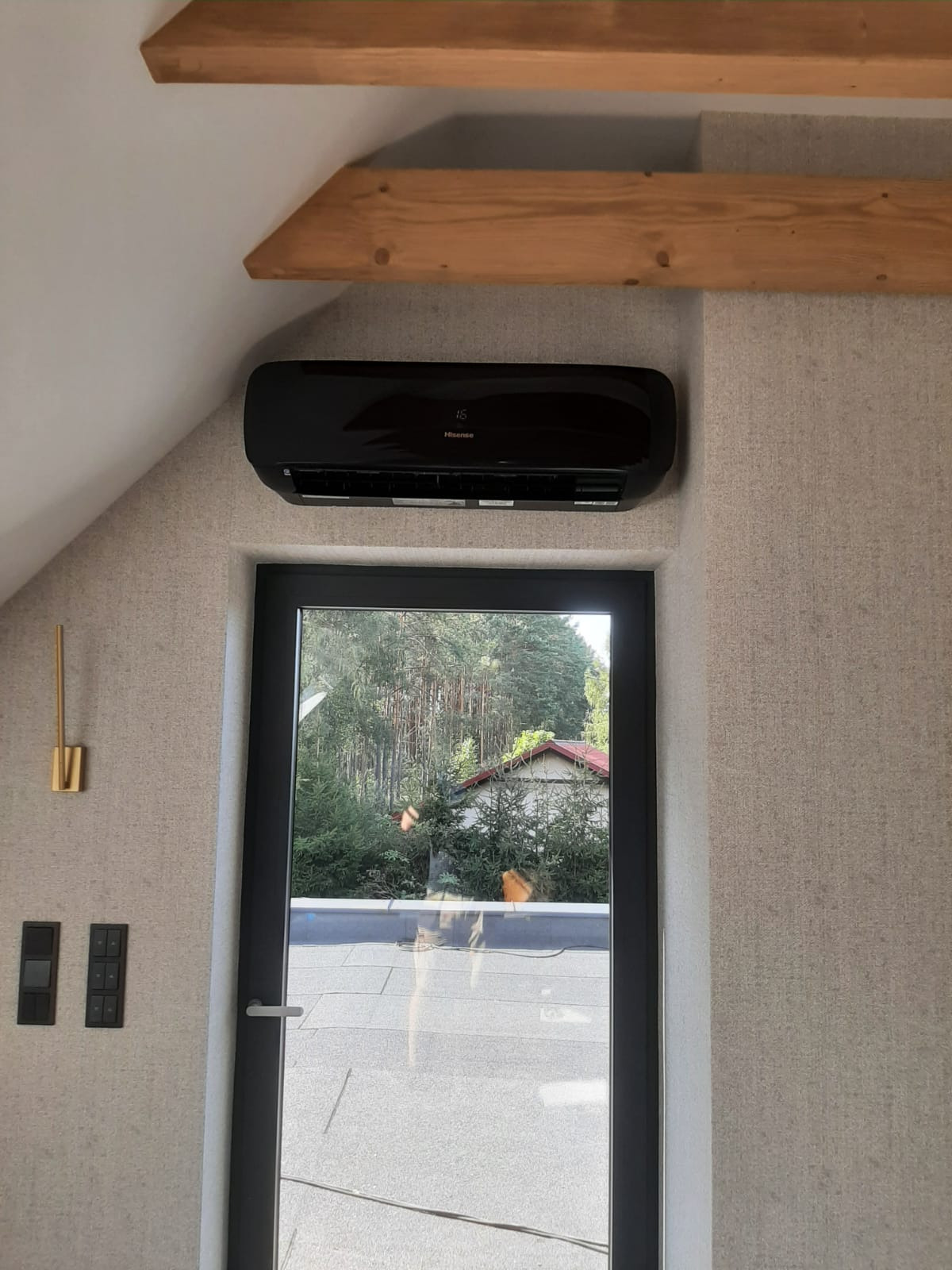 Czarna jednostka wewnętrzna klimatyzacji marki Hisense zamontowana nad oknem w pokoju na poddaszu z drewnianymi belkami stropowymi.