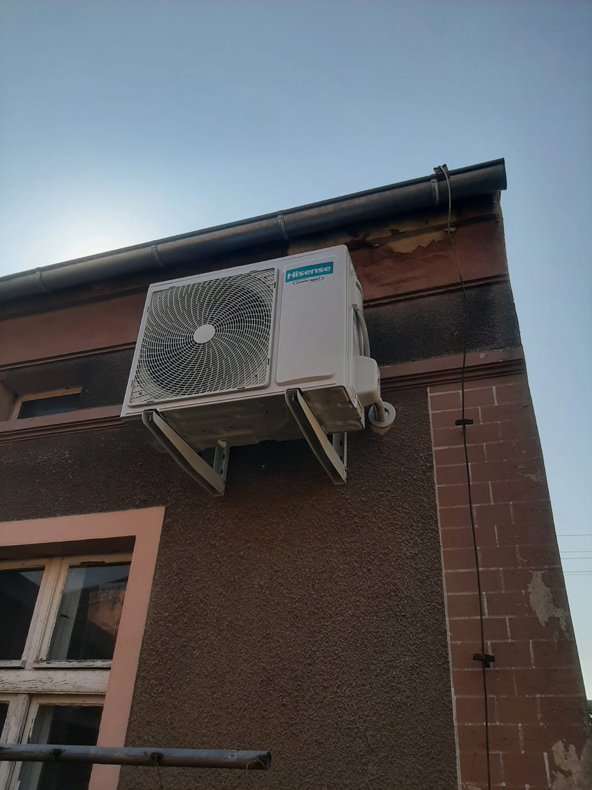 Zewnętrzna jednostka klimatyzacji Hisense zamontowana na wspornikach na elewacji budynku z widocznym oknem i rynną pod błękitnym niebem.