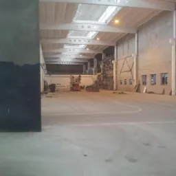 Wnętrze hali przemysłowej w trakcie prac wykończeniowych, widoczne rusztowania, podnośnik i surowe ściany przygotowywane do dalszej obróbki.