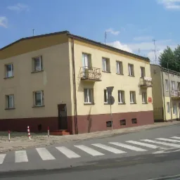 Dwupiętrowy budynek pomalowany na żółto z brązowym pasem na dole, widok z ulicy na przejście dla pieszych.