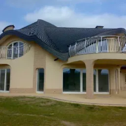 Dwupiętrowy dom jednorodzinny z zaokrąglonym dachem pokrytym ciemną dachówką, balkonami z metalową balustradą i elewacją w odcieniach beżu i cegły.