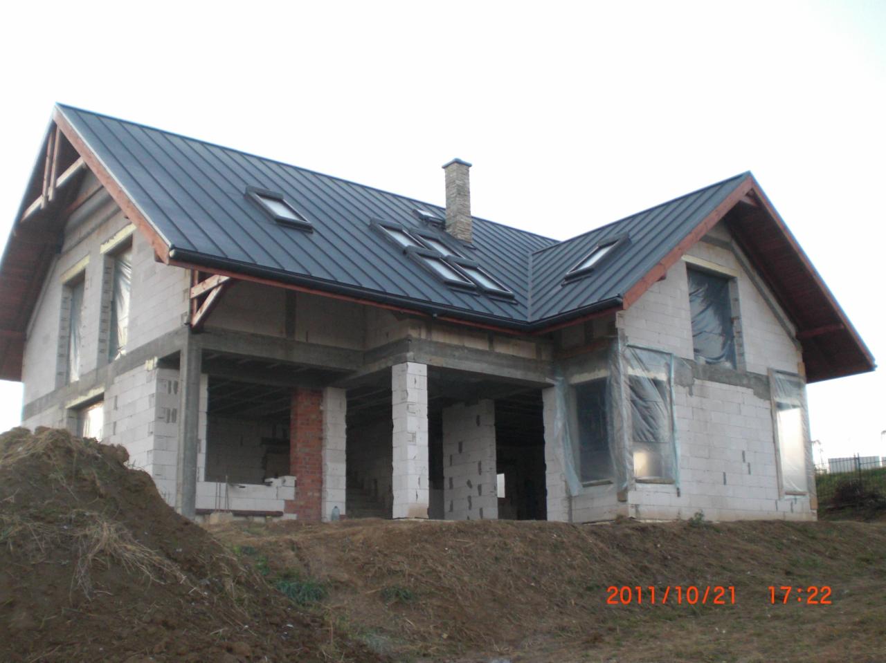 Dom w trakcie budowy z zamontowaną, ciemną blachodachówką i oknami dachowymi; widoczny komin i niezabudowane otwory okienne zabezpieczone folią.