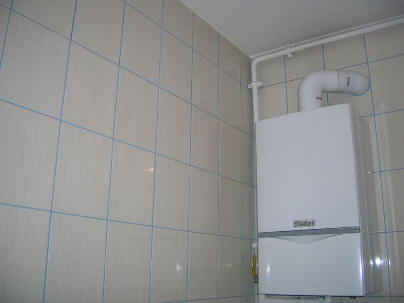 Biały piec gazowy Vaillant zamontowany na ścianie wyłożonej jasnymi, kwadratowymi płytkami z niebieską fugą w łazience.