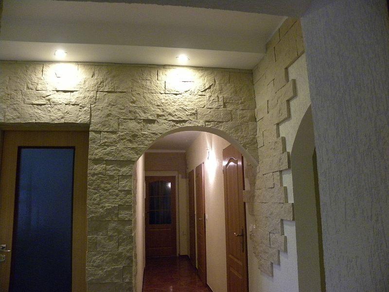 Korytarz z dekoracyjną ścianą z kamienia imitującego cegłę wokół drzwi i łuku, oświetlony punktowo, z widocznymi elementami stelaża pod sufitem.