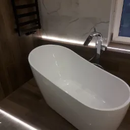 Nowoczesna, wolnostojąca wanna z chromowaną baterią, podświetlona listwą LED przy podłodze i pod oknem, na tle marmurowej ściany i drewnopodobnej okładziny.