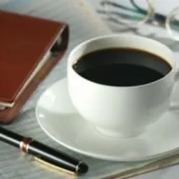 Filiżanka czarnej kawy na białym spodku, obok skórzany notatnik, pióro i okulary na gazecie, sugerujące przerwę w pracy biurowej.