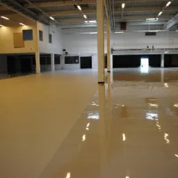 Duża hala przemysłowa z częściowo wykonaną, dwukolorową posadzką. Lewa strona matowa, beżowa, prawa strona błyszcząca, w odcieniu miodowym, z widocznymi refleksami świetlnymi.