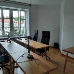 Jasne biuro z drewnianymi biurkami i czarnymi krzesłami obrotowymi, widok na budynki przez okno, lampka na biurku.