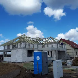 Wznoszenie konstrukcji dachu na surowym, dwukondygnacyjnym budynku z białego bloczka, z widocznymi elementami deskowania i pracującym robotnikiem, na tle błękitnego nieba z białymi obłokami.