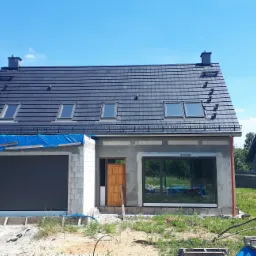 Niedokończony dom jednorodzinny z szarej cegły z ciemnym dachem, trzema oknami dachowymi i dwoma kominami, z garażem częściowo osłoniętym niebieską plandeką i brązowymi drzwiami wejściowymi,...