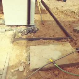 Przygotowanie terenu pod nawierzchnię: ułożone obrzeża, płyty betonowe, stos kostki brukowej, łopata i wąż ogrodowy na piaszczystym podłożu.