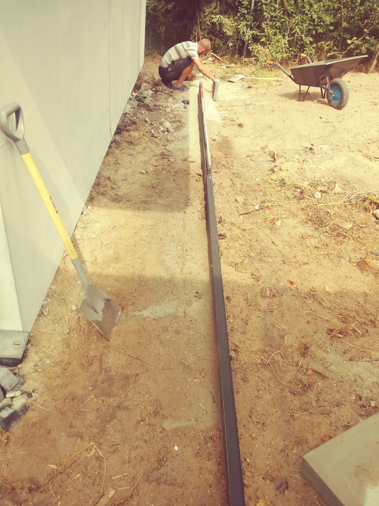 Pracownik wyrównuje poziom gruntu przy użyciu poziomicy i kielni, przygotowując podłoże pod układanie nawierzchni, obok stoi łopata i taczka.