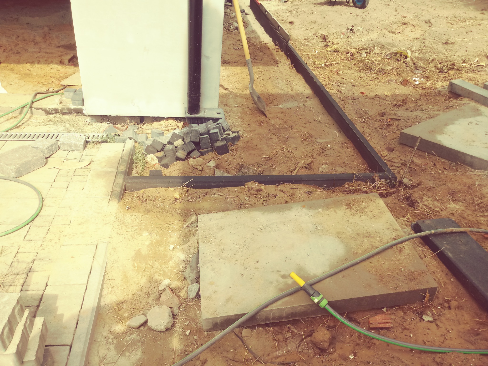 Przygotowanie terenu pod nawierzchnię: ułożone obrzeża, płyty betonowe, stos kostki brukowej, łopata i wąż ogrodowy na piaszczystym podłożu.