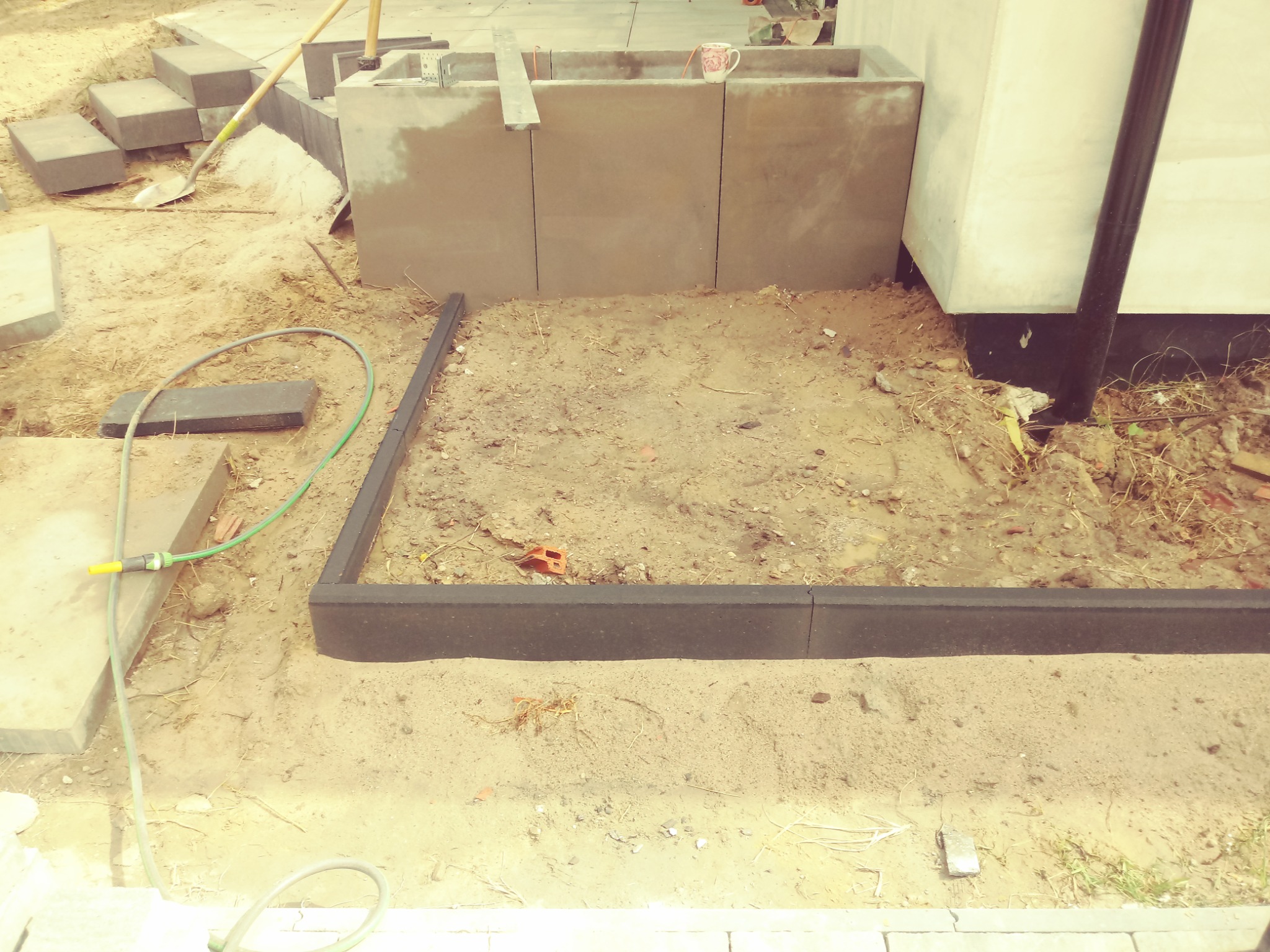 Przygotowanie terenu pod kostkę brukową: czarne obrzeża ułożone w kwadrat, wykopana i wyrównana ziemia, w tle stos kostki brukowej i szpadel.