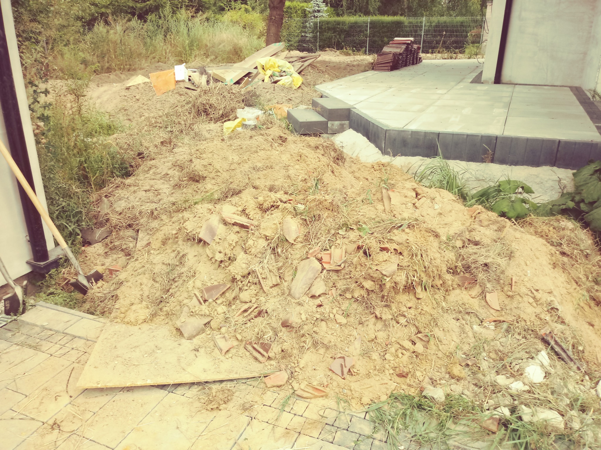 Przygotowanie terenu pod kostkę brukową w Poznaniu: Ułożone płyty tarasowe sąsiadują z pryzmą ziemi i rozrzuconymi fragmentami gruzu. W tle widoczne materiały budowlane i zieleń.