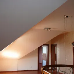 Poddasze z widocznymi sufitami podwieszanymi, skośny sufit pomalowany na szaro, na drugim planie drewniane drzwi i schody z drewnianą barierką.