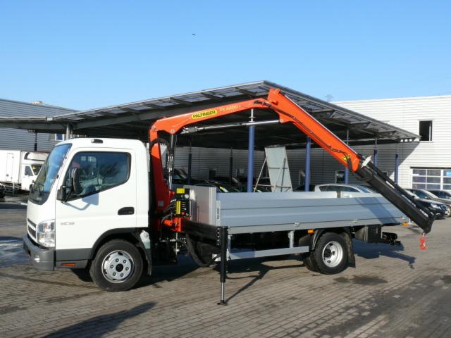 Biała ciężarówka z pomarańczowym dźwigiem Palfinger PK 8500 T, zaparkowana na zewnątrz, z wysuniętą podporą stabilizującą, na tle zadaszenia i budynku.