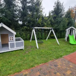 Plac zabaw w Gdańsku z drewnianym domkiem w odcieniach szarości i bieli, huśtawką z okrągłym siedziskiem oraz domkiem ze zjeżdżalnią na zielonym trawniku.