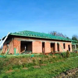 Surowy stan domu jednorodzinnego z czerwonej cegły, z zielonym dachem w trakcie budowy, widoczne rusztowania i przygotowany teren.