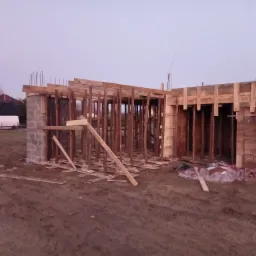Wczesny etap budowy domu z widocznym szalunkiem fundamentów i ścian, częściowo z bloczków betonowych i pustaków ceramicznych, na tle zachodzącego słońca.