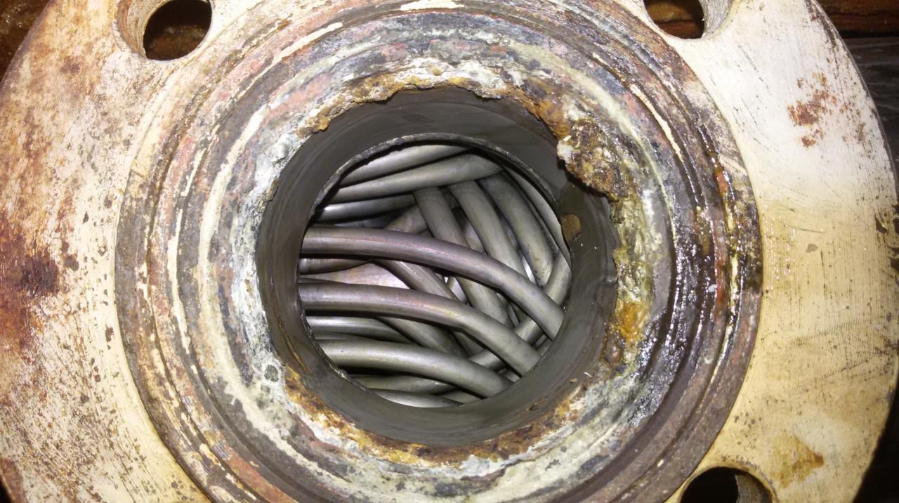 Zardzewiała flansza z widocznymi, poskręcanymi rurkami wewnątrz. Widoczne ślady korozji i osady na metalu. Specyficzne oświetlenie podkreśla fakturę.
