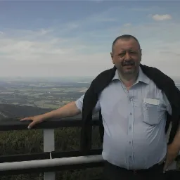 Mężczyzna z wąsami, oparty o barierkę na platformie widokowej, rozległy krajobraz w tle, pochmurne niebo.