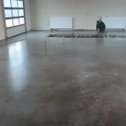 Wnętrze hali z gładką, szarą posadzką i pracownikiem w oddali, przygotowującym otwór w podłodze do dalszych prac wykończeniowych.