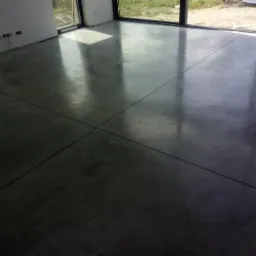 Wypolerowana, szara posadzka z widocznymi liniami podziału, odbijająca światło z dużych okien w surowym wnętrzu.