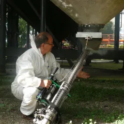 Pracownik w kombinezonie ochronnym wykonuje gazowanie przy użyciu specjalistycznego urządzenia rozpylającego, skierowanego w stronę konstrukcji przemysłowej.