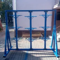 Niebieski, metalowy stojak na kółkach z trzema rzędami poziomych ramion, wykonany na zamówienie, ustawiony na brukowanym dziedzińcu przed budynkiem.
