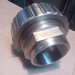 Metalowa nakrętka z gwintem wewnętrznym i sześciokątną podstawą, sfotografowana z bliska na kartonowym podłożu, widoczne ślady obróbki.